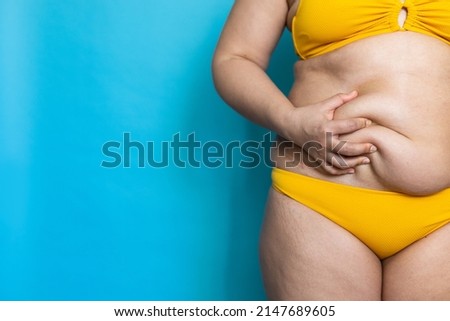 Massage woman sagging belly with hand closeup, folds on stomach, loose skin and cellulite. Overweight girl on blue background in yellow underwear, free copy space. Dieting and weight problems.