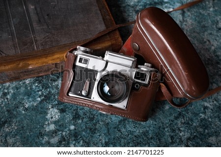 A vintage Soviet camera in a brown leather case lies on a textured blue tabletop. Nearby is an old book.