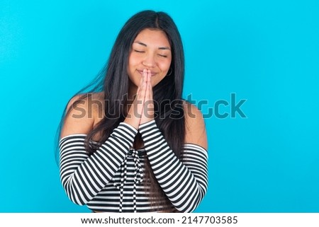 Indoor closeup of young latin woman wearing fashion clothing over blue background practicing yoga and meditation, holding palms together in namaste, looking calm, relaxed and peaceful.
