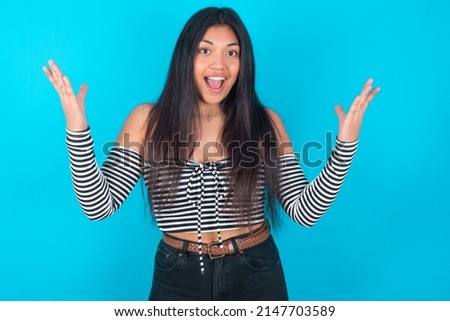 young latin woman wearing fashion clothing over blue background raising hands up, having eyes full of happiness rejoicing his great achievements. Achievement, success concept.