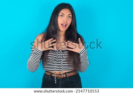 young latin woman wearing fashion clothing over blue background keeps hands on chest feeling shocked and scared, mouth widely opened, stares at camera saying: Who, me?