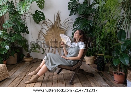 Happy young caucasian woman florist relax in home garden breathing fresh air hold paper fan in hand. Relaxed carefree millennial female gardener refreshing in indoor orangery during hot summer day Royalty-Free Stock Photo #2147712317
