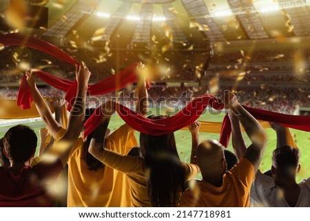 Win, victory of favourite team. Back view of football, soccer fans cheering their team with red scarfs at crowded stadium at evening time. Concept of sport, cup, world, event, competition