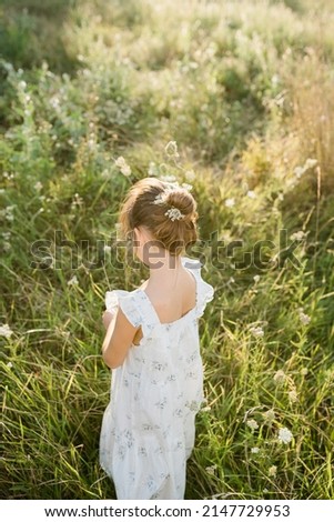 Tender little girl wearing natural white dress with wildflower motiv with wild carrot flowers, Queen Anne's lace, bird's nest, bishop's lace in hair standing in the field at summer, backlit photo