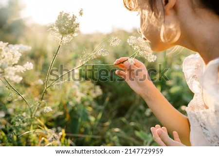Adorable little girl wearing natural white dress with wildflower motiv in green field with wild carrot flowers at summer, Queen Anne's lace, bird's nest, bishop's lace. Looks like touching the light