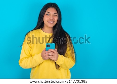 Pleased Young latin woman wearing yellow sweatshirt over blue background using self phone and looking and winking at the camera. Flirt and coquettish concept.