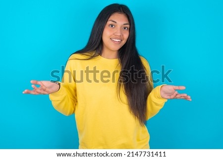 So what? Portrait of arrogant Young latin woman wearing yellow sweatshirt over blue background shrugging hands sideways smiling gasping indifferent, telling something obvious. Royalty-Free Stock Photo #2147731411