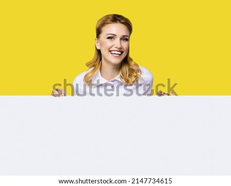 Very happy excited, laughing business woman in white confident clothing standing behind empty banner signboard with copy space text area. Success and advertising concept. Yellow background.