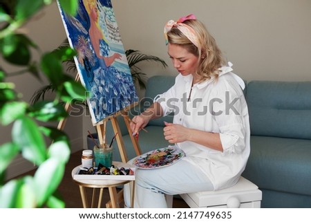 Female artist squeezing acrylic paints on palette while sitting at home or art studio, painting picture on canvas