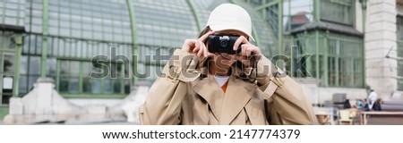 young woman in beige trench coat and baseball cap holding vintage camera, banner