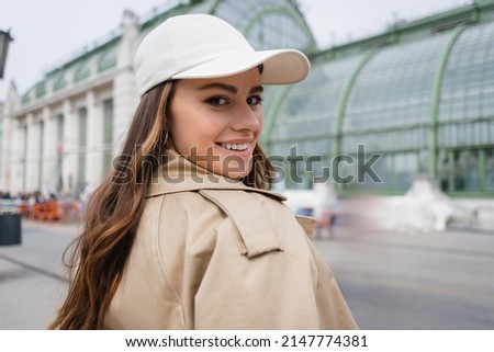 happy young woman in beige trench coat and baseball cap