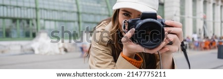 young photographer in trench coat and baseball cap taking photo on digital camera, banner
