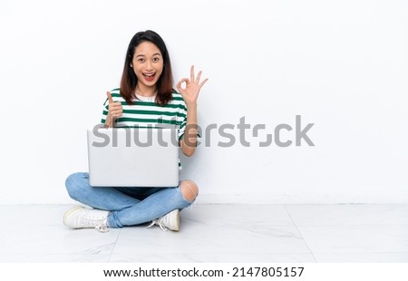 Young Vietnamese woman with a laptop sitting on the floor isolated on white wall showing ok sign and thumb up gesture