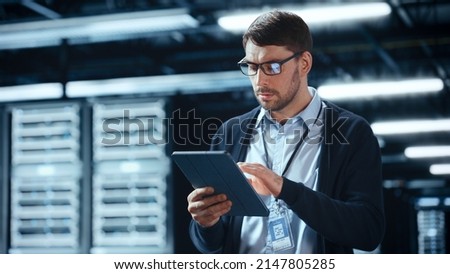 Male IT Specialist Walks Between Row of Operational Server Racks in Data Center. Engineer Uses Tablet Computer for Maintenance. Concept for Cloud Computing, Artificial Intelligence, Cybersecurity. Royalty-Free Stock Photo #2147805285