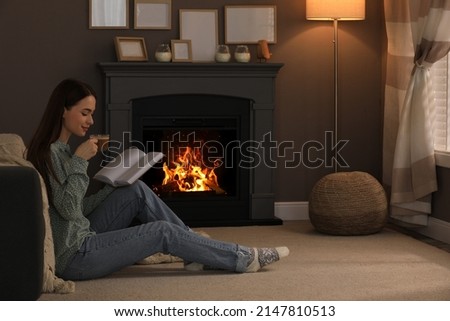 Beautiful young woman with cup of coffee reading book on floor near fireplace at home. Space for text