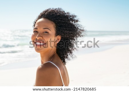 Portrait of young african american woman with curly hair at beach. Smiling black woman walking at the seaside while looking at camera. Happy carefree girl at sea. Royalty-Free Stock Photo #2147820685