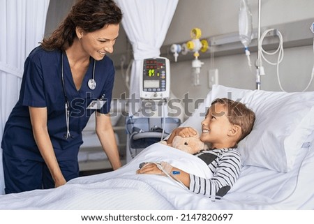 Happy young boy lying on gurney talking to friendly nurse. Smiling healthcare worker taking care of cute little boy lying on bed in hospital with IV drip. Paramedic tuck the covers back to the patient Royalty-Free Stock Photo #2147820697