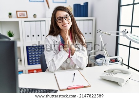 Young doctor woman wearing doctor uniform and stethoscope at the clinic praying with hands together asking for forgiveness smiling confident. 