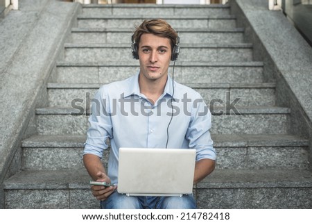 young model handsome blonde man with notebook smartphone and headphones in the city