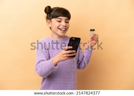 Little caucasian girl isolated on beige background buying with the mobile with a credit card