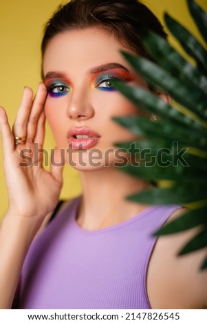 Beautiful woman with bright makeup on a yellow background in a purple sportswear with a palm leaf. Girl on vacation. photo in the style of the 80s