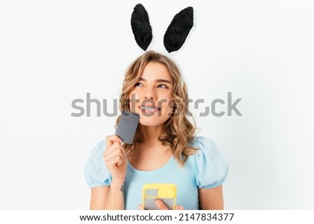Portrait happy thoughtful blonde young woman wearing Easter Bunny ears makes online shopping through her smartphone holding credit card in her hand