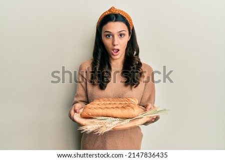 Young hispanic woman holding bread in shock face, looking skeptical and sarcastic, surprised with open mouth 