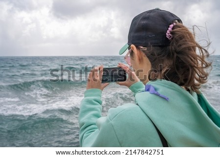 Young girl with a backpack and a smartphone takes pictures of a beautiful, stormy ocean.
