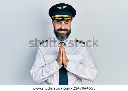 Young hispanic man wearing airplane pilot uniform praying with hands together asking for forgiveness smiling confident. 