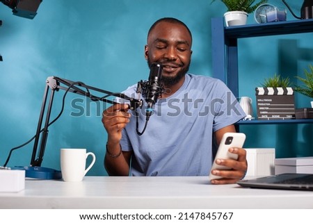 Influencer having questions and answers session using microphone in recording desk in vlogging studio. African american vlogger reading live text messages from fans looking at smartphone screen.