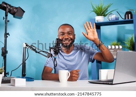 Happy vlogger waving hello at audience during online live show sitting at desk with laptop and broadcasting setup. Content creator doing greeting hand gesture in recording studio with professional