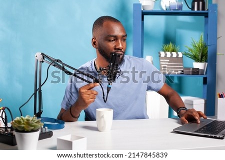 Vlogger answering questions talking at professional microphone in vlogging studio reading messages from laptop screen. Influencer giving feedback to followers in live broadcasting session.