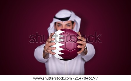 Arab man holding soccer ball in hand with Qatar flag standing on red background. Royalty-Free Stock Photo #2147846199