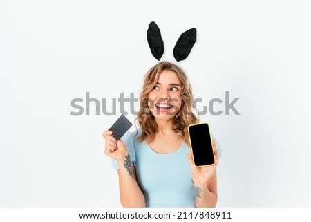 Cheerful young woman wearing Easter Bunny ears holding bank card and smartphone with blank display while standing on white background