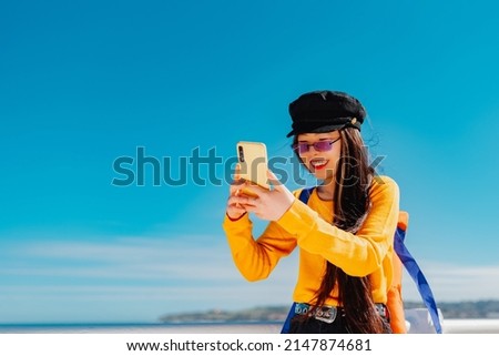 Young Asian woman wearing sunglasses, beret and a yellow sweater takes self-portraits with her smartphone. Chinese girl sightseeing