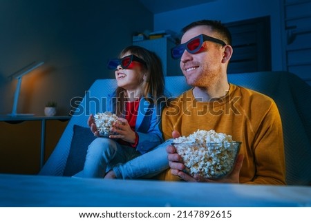 Father and daughter watching movie at home using 3D eye glasses and eating popcorn. Family activities. Selective focus on the male face. Blue light from the screen.