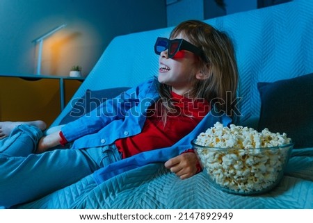 Little girl watching movie at home using stereo 3D eye glasses and eating popcorn. Selective focus. Blue light from the screen.