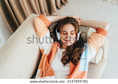 Young relaxed woman is laying down on sofa and  listening to a music with headphones on her ears.  Royalty-Free Stock Photo #2147899785