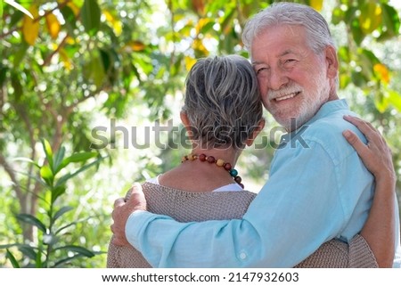 Rear view of a beautiful senior couple walking in the woods tenderly embraced while the man looks at the camera. Smiling elderly grandparents enjoy a healthy lifestyle in the public park