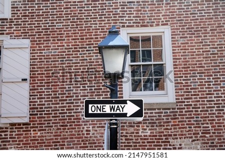 One way sign hanging on a black pole with street lamp, with a traditional brick wall on the back with white window