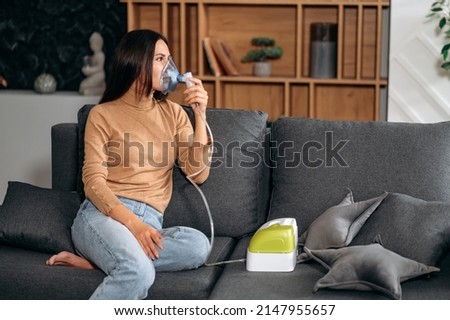 Sick upset caucasian brunette woman with an inhaler. Unhealthy female doing inhalation at home, she use nebulizer and inhaler for the treatment, sitting on the couch at home, need treatment Royalty-Free Stock Photo #2147955657