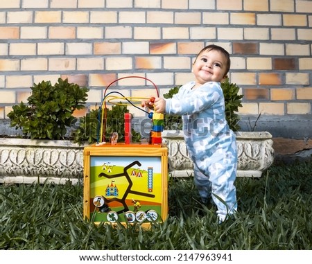Baby boy wearing a blue and white outfit sitting on a lawn with a brick wall in the background playing with a colorful toy. Mention of babies and expressions, first lessons.