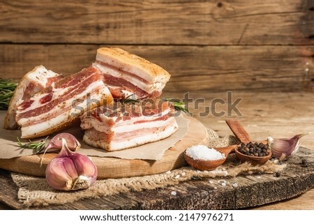 Pieces of salty high-fat meat cooked with spices. Salo, bacon, lard, silverside, gammon. Garlic, fresh rosemary, spices. Hard light, dark shadow, wooden background, copy space Royalty-Free Stock Photo #2147976271