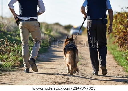 Walking along the crimescene. Rear view shot of two policeman and a dog walking down a rural road. Royalty-Free Stock Photo #2147997823