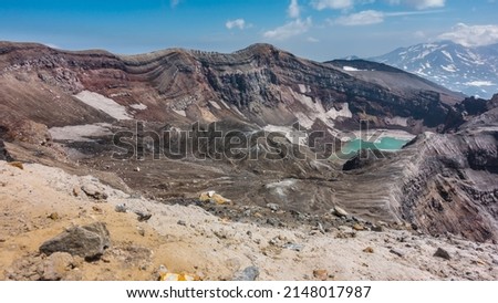 There is a turquoise acid lake on the top of an active volcano. The layered structure of the steep slopes of the crater, melted snow, stony soil is visible. Blue sky. Kamchatka. Gorely Volcano Royalty-Free Stock Photo #2148017987