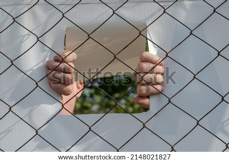 Two hands hold the banner attached to Chain-link fencing. Mature man hiding behind white banner with rectangular perforated wind hole.
