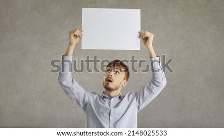 Studio portrait of man holding up paper banner with free space for text. Young guy with open mouth and surprised face expression showing empty blank mockup poster standing isolated on grey background