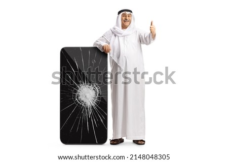 Full length portrait of an arab man in ethnic clothes with a big smarphone with cracked screen showing thumbs up isolated on white background
