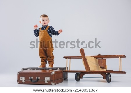 A cheerful child in a brown overalls is standing on a suitcase. Little traveler with a suitcase. Wooden plane, children's eco toys