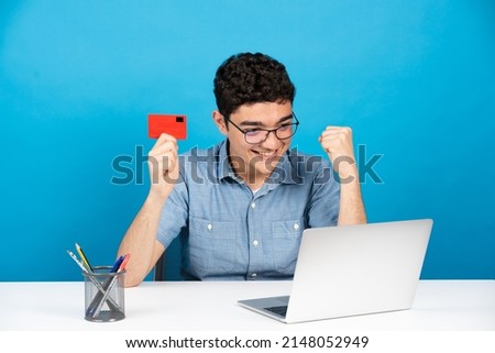 Happy hispanic guy celebrating online purchase. Teenager holding credit card in front of laptop isolated on blue background.